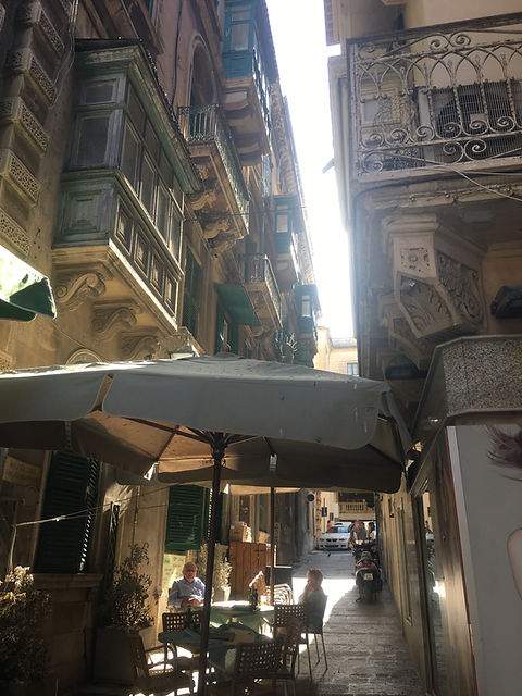 Blick durch eine enge Straße auf Malta mit Café und Sonnenschirmen.
