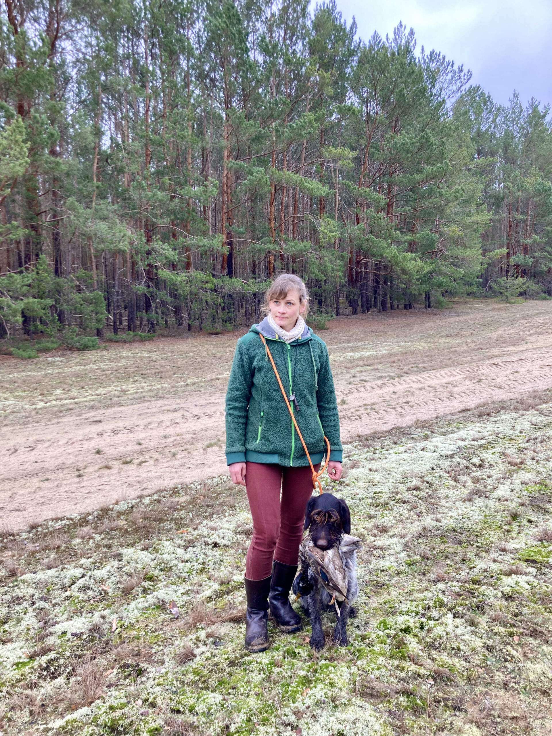 Ein Kind steht mit einem Hund auf einem Feld am Rand eines Kiefernwaldes.