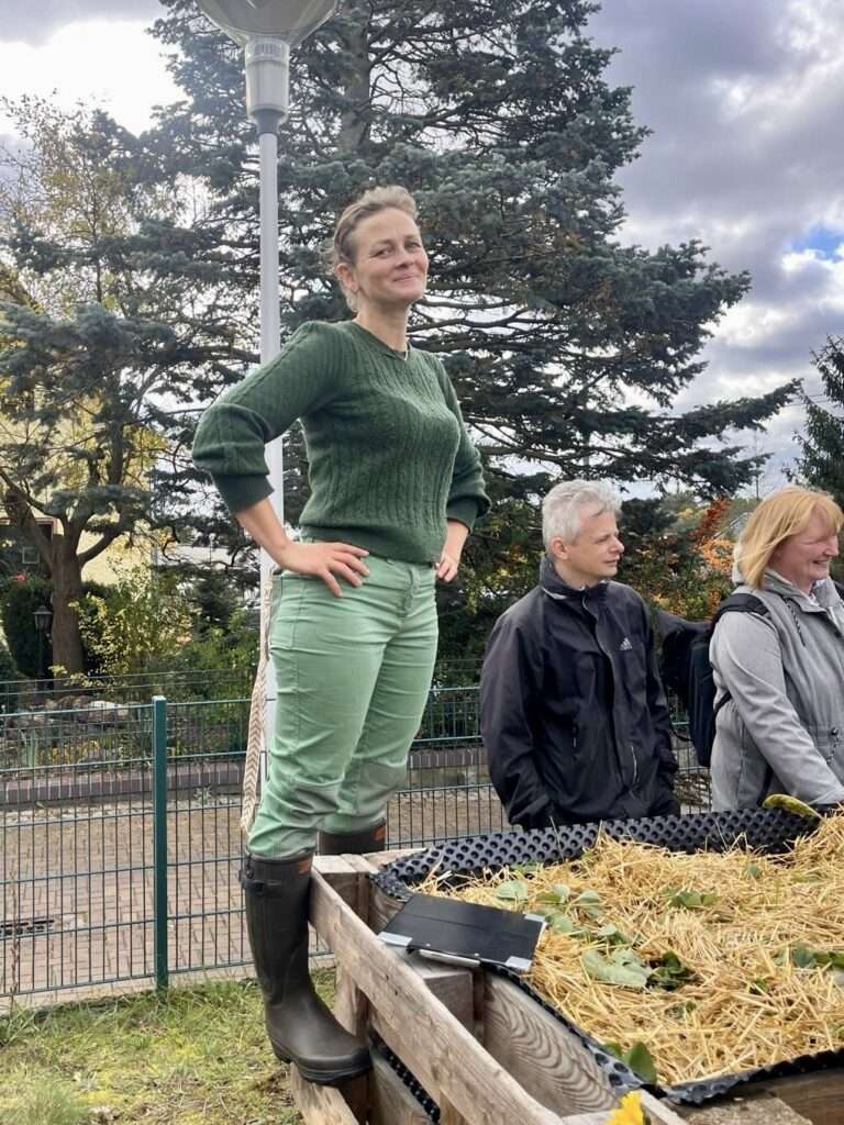 Gunhild Rudolph steht im grünen Pullover an einem Hochbeet und erklärt den Aufbau eines Komposts vor einer Gruppe.