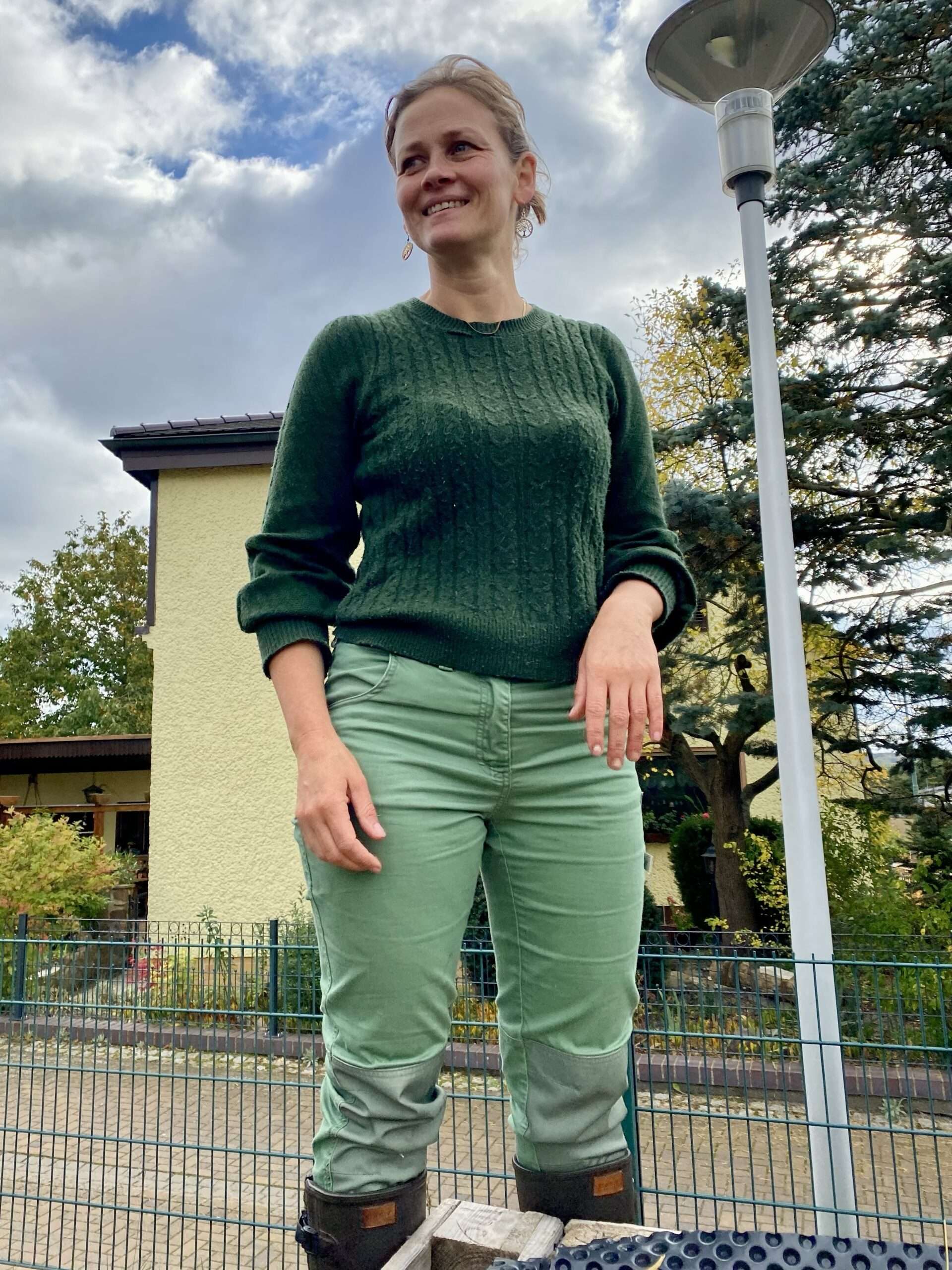 Gunhild Rudolph steht im grünen Pullover vor einem hellgelben Gebäude mit Zaun, im Hintergrund Bäume und Wolkenhimmel.